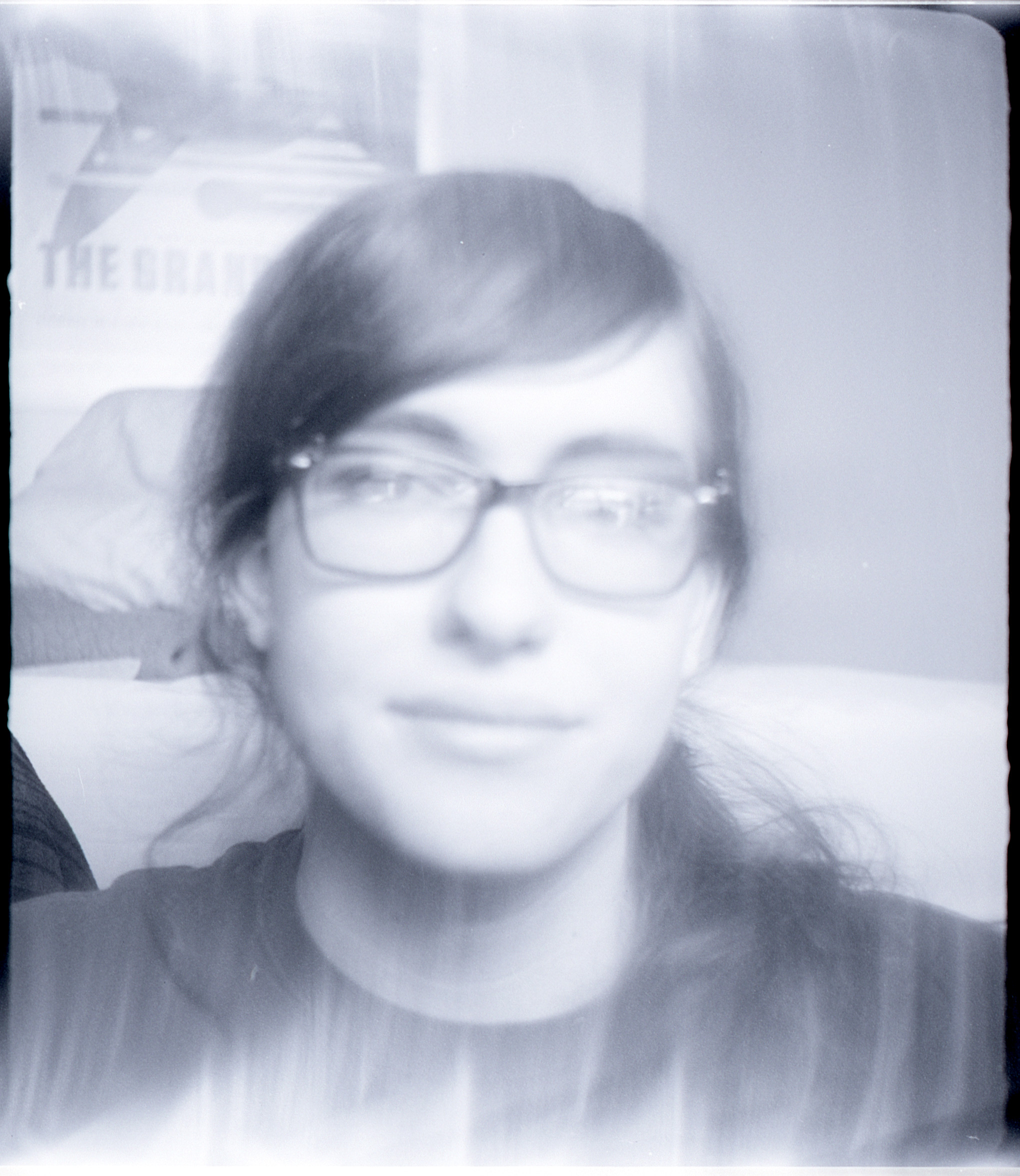 a very washed out black and white picture of a blurry girl looking at the camera. light from the window in front of her is reflected in her glasses