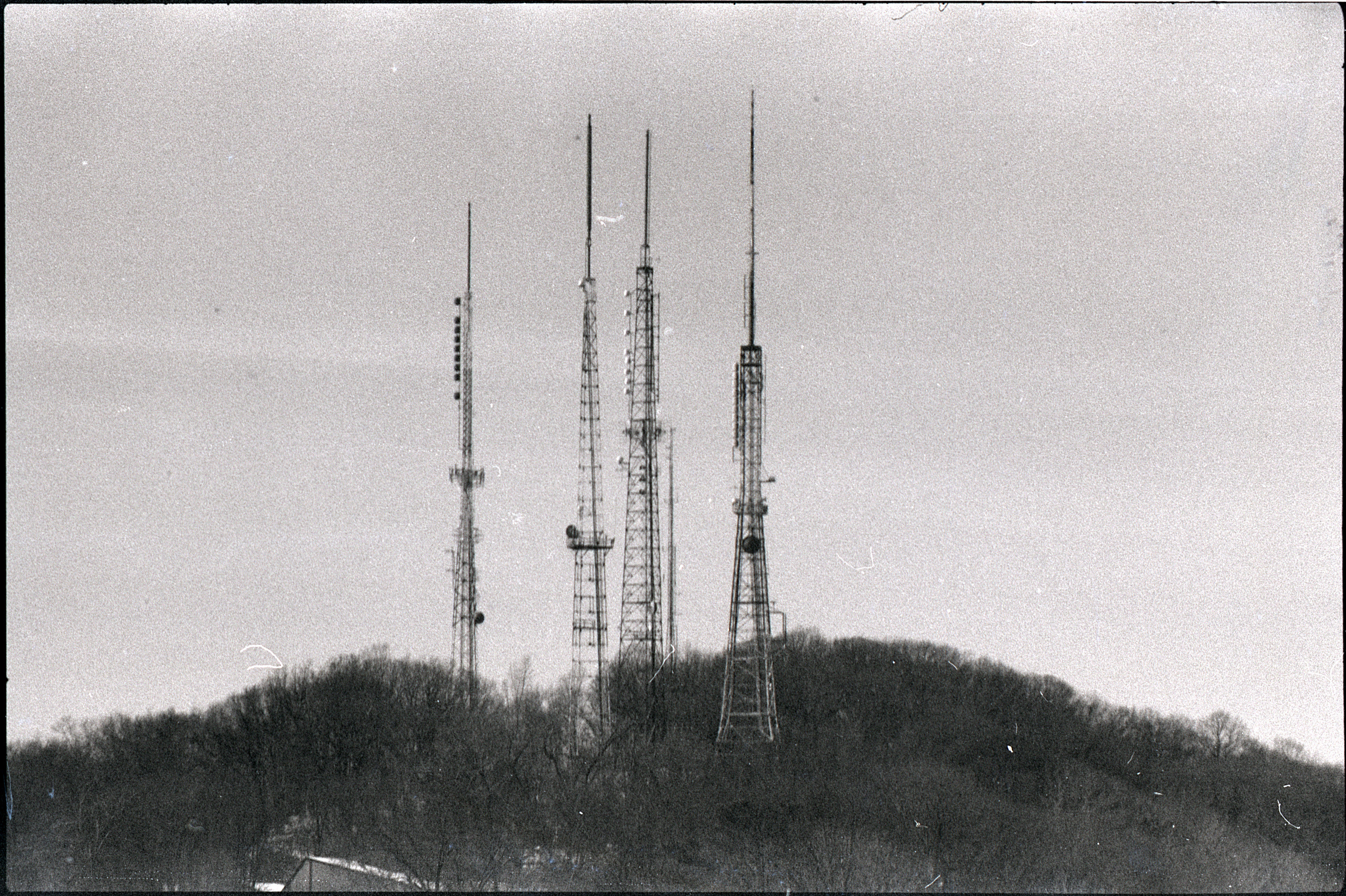 photo, black and white with some film artifacting: four radio towers covered in antennae rising above trees on a hilltop to pierce the sky