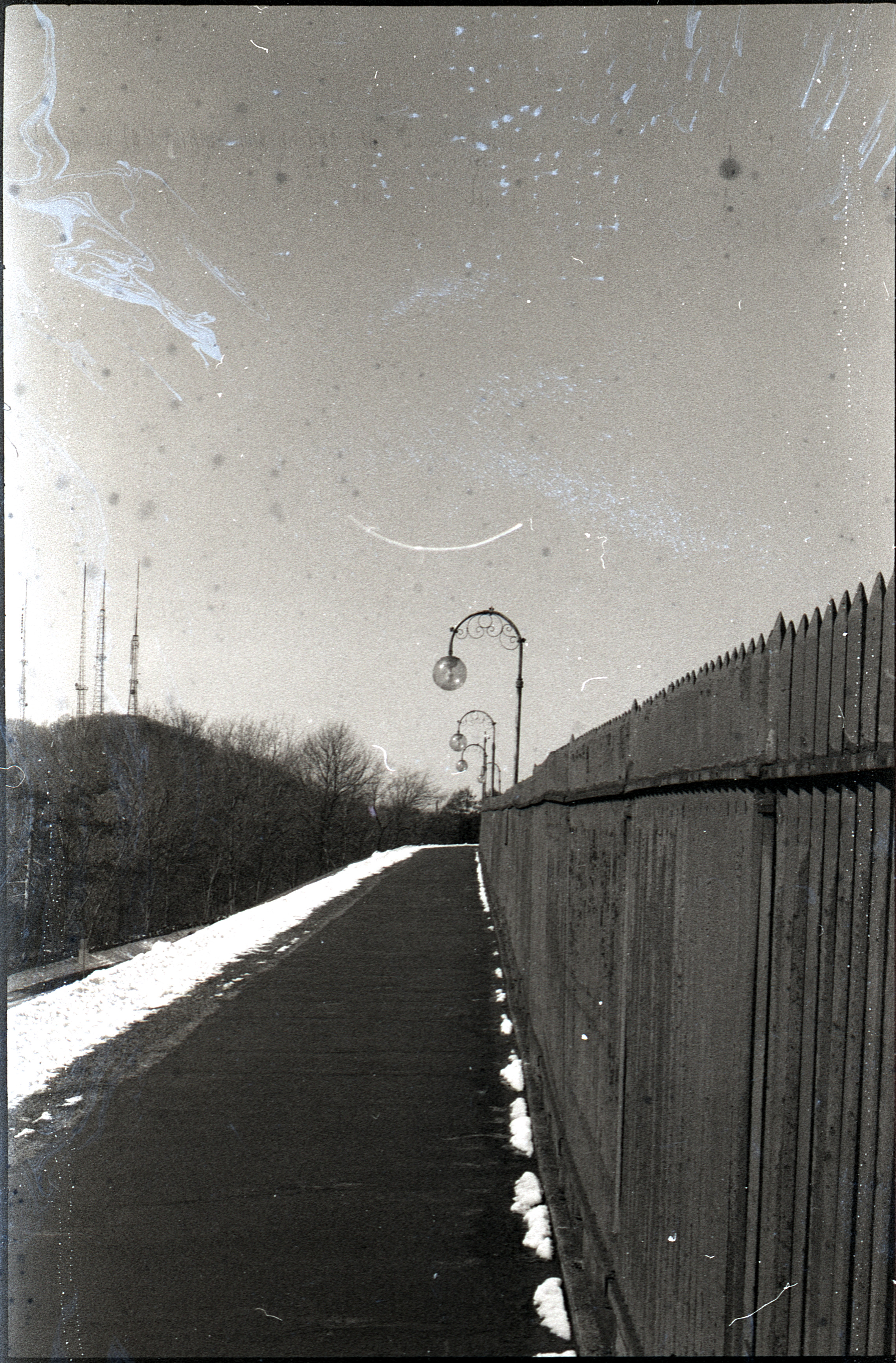 photo, black and white with significant film artifacting: a sidewalk next to a fence with spherical streetlamps hanging over it
off in the background, the hill with four radio towers is visible