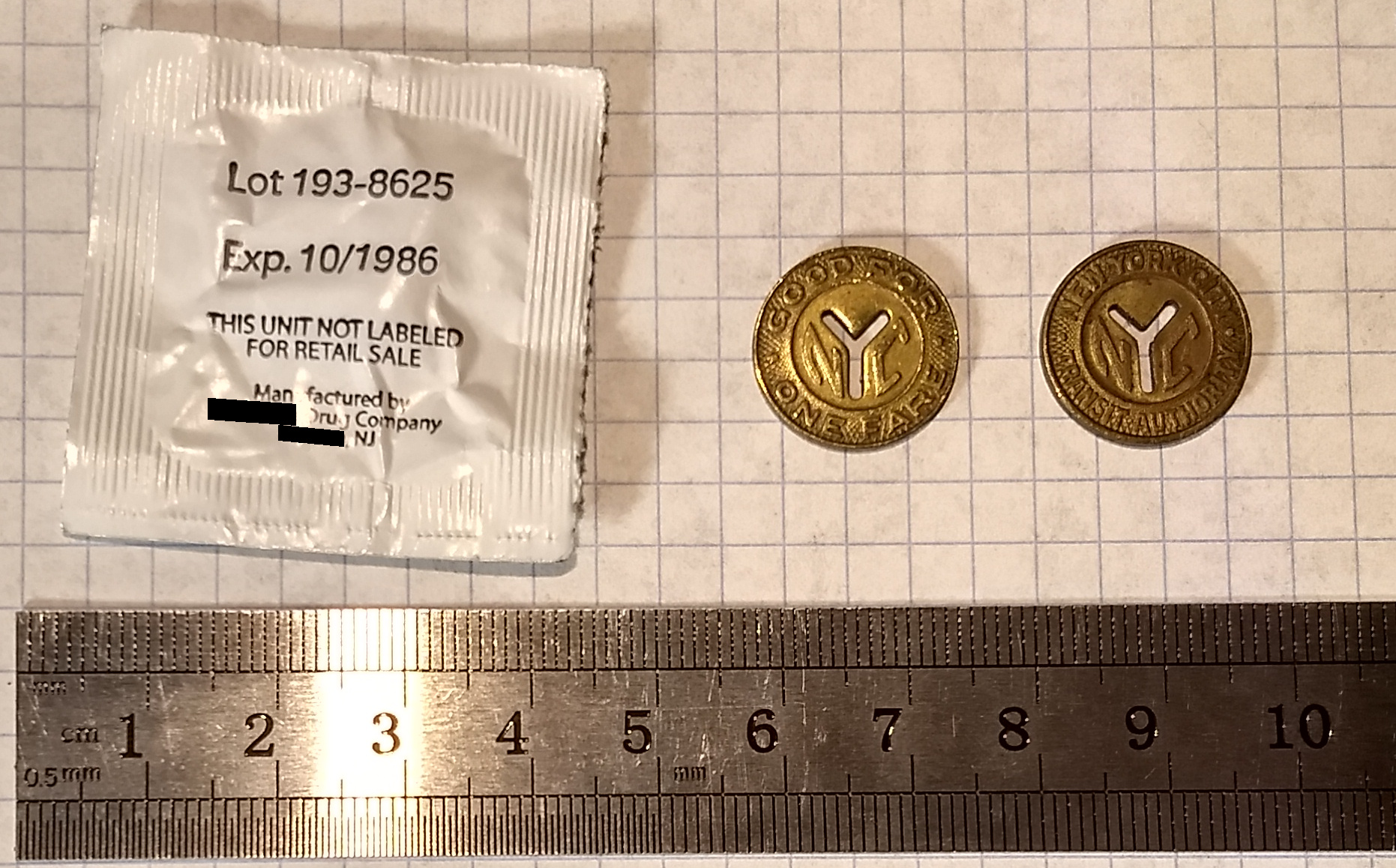 the front and back of an old NYC subway token with the letter Y cut out of it on a sheet of graph paper next to a ruler and a paper packet labeled as expiring in 10/1986